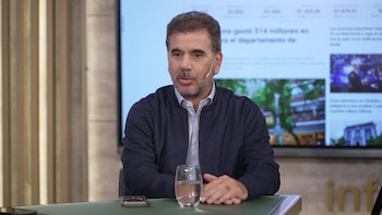 Hombre de mediana edad con barba en chaqueta azul oscuro sentado frente a una mesa verde con un vaso de agua. Una pantalla de noticias está al fondo