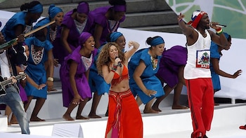 Shakira y Wyclef Jean cantando en un escenario con bailarines. Shakira viste top naranja y falda roja, Wyclef Jean lleva camiseta blanca con bandera de Haití