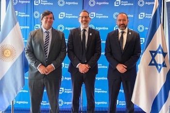 Tres hombres con traje y corbata de pie ante un fondo azul institucional, flanqueados por las banderas de Argentina e Israel