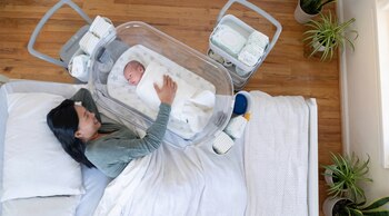 Vista aérea de mujer sonriente en cama junto a bebé en cuna transparente. Suministros para bebé apilados y plantas verdes en macetas sobre suelo de madera