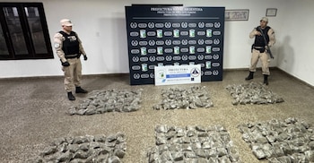 Dos oficiales de Prefectura con uniforme, uno con rifle, de pie junto a una gran cantidad de droga en paquetes sobre el suelo, con carteles