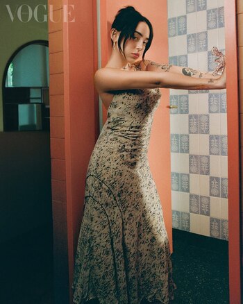 Nicki Nicole, mujer joven de cabello oscuro recogido y flequillo, lleva un vestido largo estampado, posando con brazos extendidos contra una pared rosa y azulejos