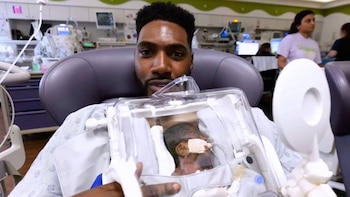 Un hombre sentado en un sillón médico sostiene una pequeña cápsula transparente con un bebé prematuro con sondas médicas
