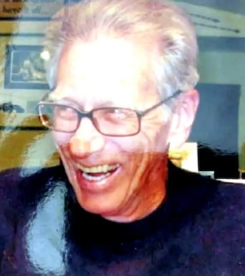 Primer plano de un hombre de cabello canoso y gafas oscuras, sonriendo y mostrando los dientes, con un suéter oscuro