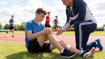 Un joven sentado en la hierba se sujeta la rodilla mientras un adulto inclinado a su lado le ofrece ayuda cerca de una pista de atletismo.