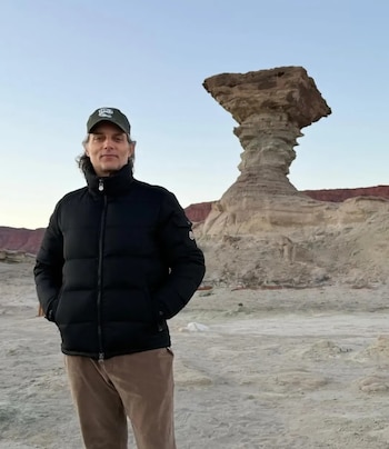 Hombre de cabello largo y campera negra posa en un paisaje árido con una formación rocosa en forma de hongo y montañas al fondo