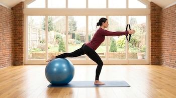 Mujer en leggings negros y camiseta granate haciendo Pilates sobre una colchoneta. Apoya una pierna en balón azul y sostiene un aro. Fondo con ventanales.