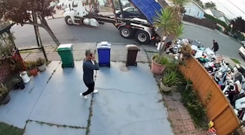 Un camión de basura descarga una pila masiva de residuos junto a una valla de madera en un jardín. Un hombre habla por teléfono cerca de tres botes de basura