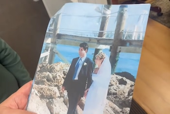 Una mano sostiene una fotografía impresa de una pareja vestida de novios caminando cerca de rocas y un muelle de madera junto al mar