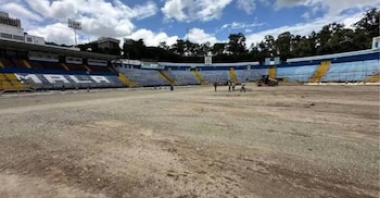 Campo de tierra en un estadio con gradas azules y amarillas. Varias personas, una máquina de construcción y árboles en el fondo