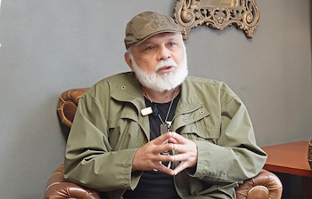 Un hombre de mediana edad con barba blanca, gorra verde oliva y chaqueta a juego está sentado en un sillón de cuero marrón, con las manos entrelazadas