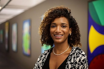 Retrato de Amanda Carneiro, mujer de cabello rizado y sonrisa amable, con collar de perlas y chaqueta estampada, delante de obras de arte abstractas difuminadas