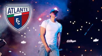 Hombre con gorra, camiseta blanca y pantalón vaquero sujetando un micrófono en un escenario con luces, y el escudo del Atlante FC.