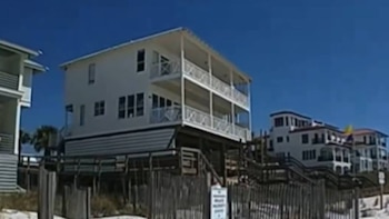 Imagen de propiedades blancas de varios pisos con balcones, elevadas sobre una zona de playa arenosa, con vallas de madera y un cielo azul claro