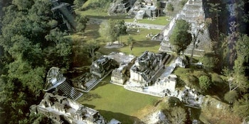 Vista aérea de las ruinas mayas de Tikal, con estructuras de piedra grises y pirámides parcialmente cubiertas por la selva tropical y zonas de césped verde