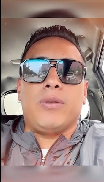 Primer plano de Christian Cueva, un hombre con cabello oscuro y gafas de sol reflectantes, vistiendo una chaqueta gris, dentro de un vehículo