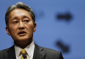 Retrato de Kazuo Hirai con traje oscuro, camisa blanca y corbata amarilla frente a un micrófono y un fondo azul