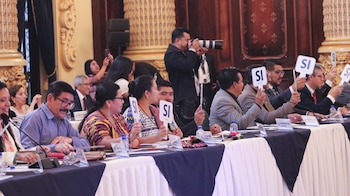 Delegados sentados en una mesa en una sala formal, levantan carteles blancos con la palabra 'SÍ'. Un fotógrafo captura la escena al fondo