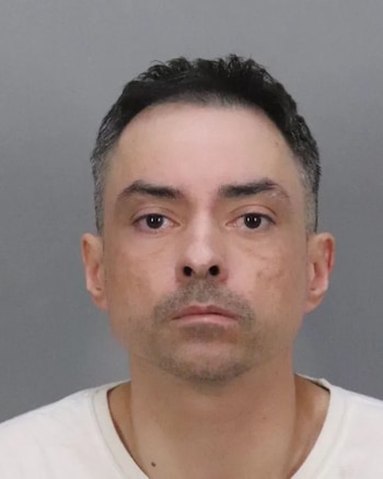 Fotografía policial de un hombre adulto con cabello corto oscuro, barba corta y camiseta blanca sobre un fondo gris