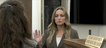 Una mujer de cabello claro y chaqueta oscura levanta su mano derecha ante una persona en la sala de un tribunal