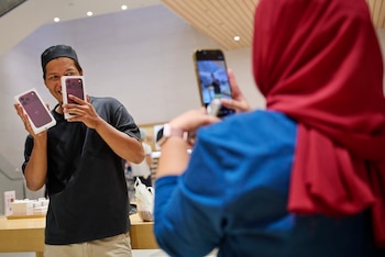 Un hombre con gorro sostiene dos cajas de iPhone frente a su rostro mientras una mujer con hiyab rojo lo fotografía con su teléfono