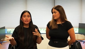 Dos mujeres vestidas con prendas oscuras permanecen de pie en una oficina frente a una ventana con persianas y escritorios con monitores