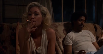 Mujer rubia sentada en un sofá, fuma un cigarrillo. Hombre con bigote y cabello rizado oscuro sentado a su derecha