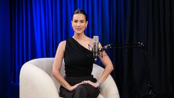 Luli Fernández sentada en un sillón blanco con un micrófono de estudio, frente a cortinas azules y negras. Viste top negro y pantalón oscuro