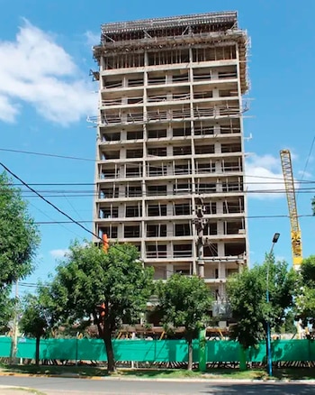 Vista aérea de un edificio de hormigón de muchos pisos sin terminar, con andamios visibles en la parte superior, rodeado de árboles y edificios bajos