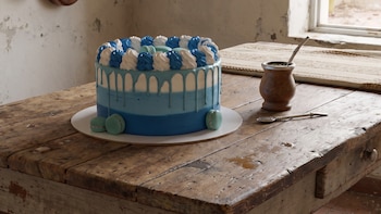 Torta con cobertura azul degradada, crema blanca y azul, y macarons celestes. Mate con bombilla de metal. Mesa de madera, cacerolas y ventana.