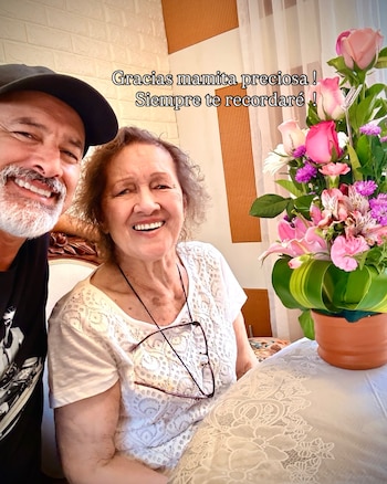 Hombre con barba gris y gorra negra sonríe junto a mujer de cabello rizado gris con lentes colgando. Arreglo floral de rosas y flores moradas. Texto