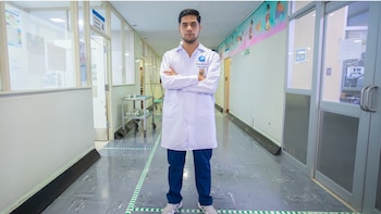Um jovem de jaleco branco e calça azul, braços cruzados, parado no corredor de um hospital. Paredes claras, janelas e carrinhos médicos são refletidos