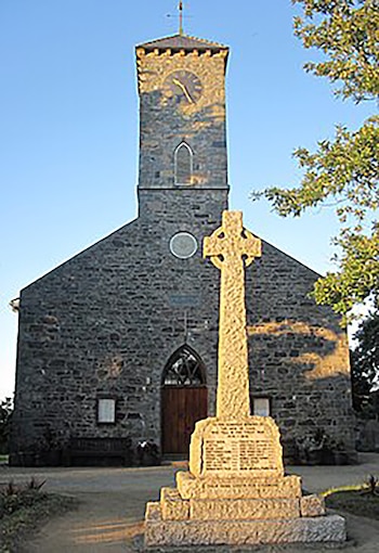 Una iglesia de piedra con torre de reloj y una puerta de madera oscura se alza bajo un cielo azul claro, con una gran cruz celta de piedra en primer plano