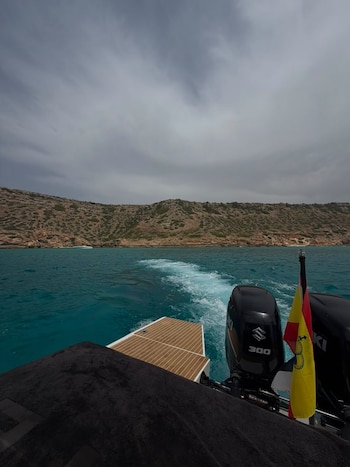 Vista de la popa de una embarcación con dos motores fuera de borda, bandera de las Islas Baleares, agua turquesa con estela, costa rocosa y cielo nublado