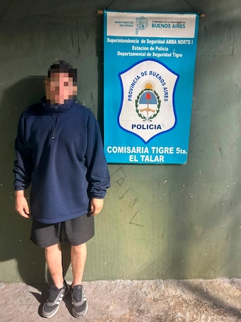 Hombre con rostro pixelado vestido de azul y negro de pie frente a pared verde y cartel azul de la Policía de la Provincia de Buenos Aires
