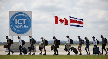 Una fila de turistas camina por una pista de aeropuerto, llevando equipaje. Al fondo, un letrero azul del ICT y las banderas de Canadá y Costa Rica ondean bajo un cielo nublado.