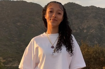 Una joven con cabello oscuro rizado y tez clara, viste una camiseta blanca y dos collares, posa sonriendo ante montañas con vegetación