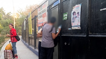 Una mujer con bolsas y personas ante un portón negro con avisos pegados sobre vacunación contra el sarampión y clases escolares.