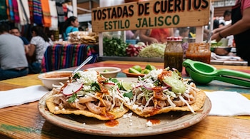 Dos tostadas de maíz con cueritos, repollo, rábano, queso y salsa sobre un plato, con un letrero de madera de fondo.