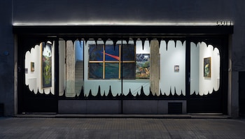 Fachada de vidrio de una galería de arte con un marco negro en forma de dientes y varias pinturas expuestas en paredes blancas