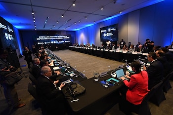 Sala de conferencias con mesa grande en forma de U, personas sentadas, pantallas con logos, laptops, agua y micrófonos en la mesa