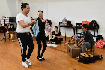 Un hombre y una mujer bailan con pañuelos, mientras otros jóvenes tocan instrumentos musicales tradicionales como guitarra y cajón en un salón con piso de madera