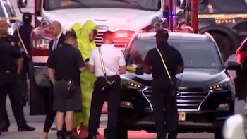 Un grupo de personas, algunos vestidos de oficiales, rodea un SUV negro. Una persona con traje de protección amarillo y vehículos de emergencia visibles
