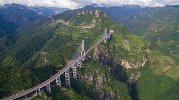 Vista aérea de un puente atirantado largo con dos torres y múltiples cables, que atraviesa un valle verde rodeado de montañas y un túnel en uno de sus extremos