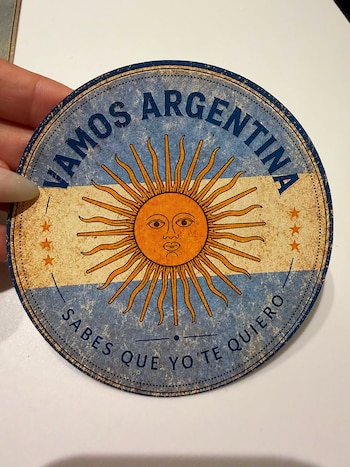 Una mano sostiene un posavasos redondo con diseño de la bandera argentina, Sol de Mayo, cuatro estrellas, y las frases 'VAMOS ARGENTINA' y 'SABES QUE YO TE QUIERO'