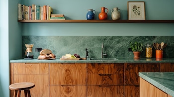 Cocina con encimera de mármol verde, gabinetes de madera, estante con libros y jarrones, lavabo, cafetera, pan, queso, uvas, planta, jarra y utensilios.