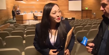 Mujer asiática, Eileen Wang, con chaqueta azul oscuro y labios rojos, habla con un reportero que sostiene un micrófono de CBS LA en una sala con sillas vacías
