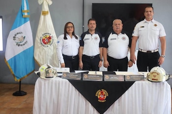 Cuatro personas uniformadas de bomberos. Dos banderas: de Guatemala y de Bomberos Voluntarios. Dos cascos de bombero sobre una mesa blanca