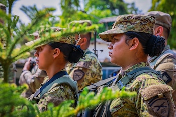 Dos mujeres militares con uniforme de camuflaje verde, gorra y equipo, con el cabello recogido, se ven de perfil en un entorno exterior frondoso