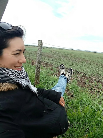 Mujer sonriente con abrigo negro y bufanda a cuadros sentada cerca de un cerco y un campo de cultivo